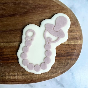 May include: A white cookie with a pink outline of a pacifier and a beaded necklace. The cookie is on a wooden surface.
