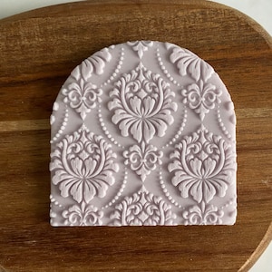 May include: A light purple silicone cake decorating stamp with a damask pattern. The stamp is shaped like an arch.