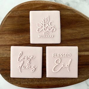 May include: Three pink square cookies with white lettering. The top cookie says "Eid Mubarak" in Arabic script and English. The left cookie says "love & duas". The right cookie says "Blessed Eid".