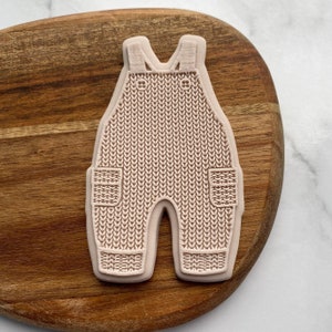 Könnte beinhalten: Eine rosa Silikonform in Form einer gestrickten Latzhose mit Taschen. Die Form ist zum Backen und Dekorieren gedacht.