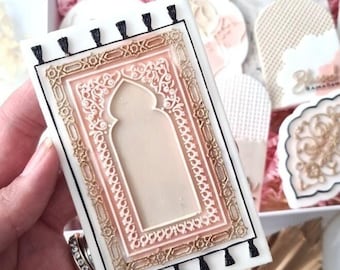 May include: A white and pink decorated cookie shaped like a prayer rug with a gold border and black tassels. The cookie is in a box with other decorated cookies.