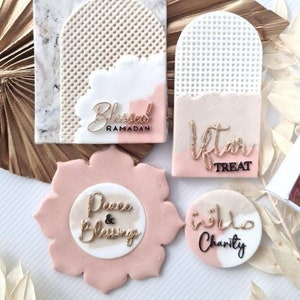 May include: Four round, decorated cake toppers with different designs and text. The toppers are decorated with white, pink, and gold icing. The text on the toppers reads "Blessed Ramadan", "Iftar Treat", "Peace & Blessings", and "Charity".