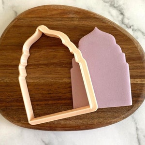 Può includere: Un tagliapasta in plastica rosa chiaro a forma di arco con la parte superiore a punta. Il tagliapasta è su una superficie di legno.