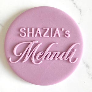 The image shows a personalized fondant embosser or stamp, for creating custom decorations on baked goods for a Mehndi ceremony. It features the custom text above the word "Mehndi" in a script font. These embossers are used to imprint designs and text onto fondant, icing, or dough, adding a personalized touch to cakes, cupcakes, and cookies for special occasions like weddings and Mehndi celebrations.