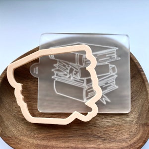 May include: A clear acrylic stamp with a design of a stack of books. The stamp is on top of a wooden cookie cutter in the shape of a book.
