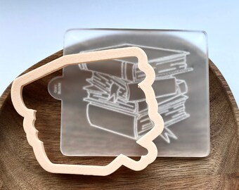 May include: A clear acrylic stamp with a design of a stack of books. The stamp is on top of a wooden cookie cutter in the shape of a book.