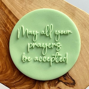 Puede incluir: Un sello redondo verde con el texto "May all your prayers be accepted" en letras blancas cursivas.