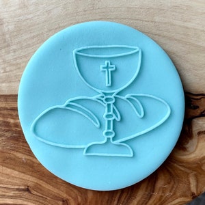 May include: A light blue silicone cookie cutter shaped like a chalice with a cross and a loaf of bread.