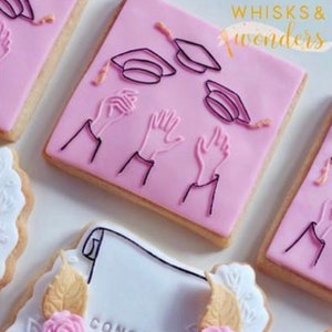 May include: Pink and white decorated sugar cookies with graduation cap and hand designs. One cookie has the text "CONGRATS" on it.