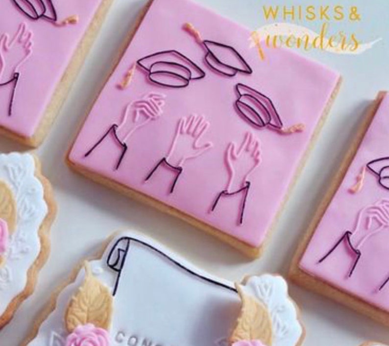 May include: Pink and white decorated sugar cookies with graduation cap and hand designs. One cookie has the text "CONGRATS" on it.