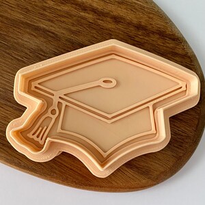 May include: A beige plastic cookie cutter shaped like a graduation cap. The cap has a tassel and a detailed outline.