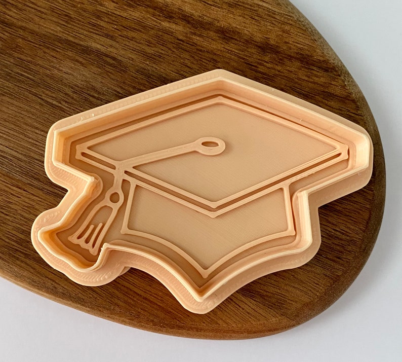 May include: A beige plastic cookie cutter shaped like a graduation cap. The cap has a tassel and a detailed outline.