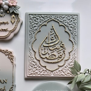 Peut inclure: Une plaque décorative carrée avec un fond dégradé vert clair et rose. La plaque présente un motif de calligraphie arabe de couleur or dans un cadre blanc orné.