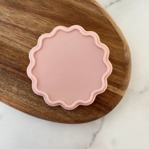 May include: A pink, scalloped, round coaster. The coaster is made of a smooth, soft material and has a slightly raised edge.