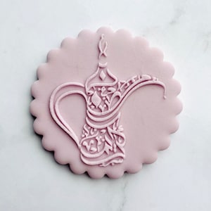Puede incluir: Una galleta redonda de color rosa con un diseño en relieve detallado de una cafetera árabe tradicional. El diseño presenta una caligrafía intrincada y motivos florales.