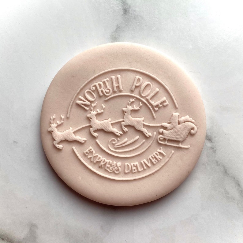 May include: A pink round silicone stamp with a raised design of three reindeer pulling a sleigh with Santa Claus. The stamp says "North Pole Express Delivery" in a circle around the reindeer and sleigh.