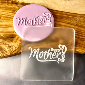Puede incluir: Un sello de acrílico transparente con el texto "Happy Mother's Day" en una fuente cursiva. El sello está sobre una superficie de madera.