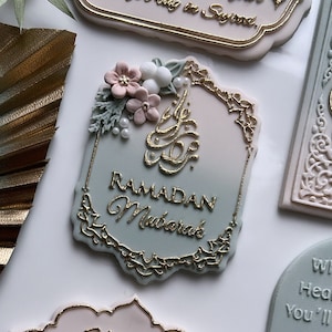Peut inclure: Une plaque décorative avec un fond dégradé bleu clair et rose. La plaque a une bordure dorée ornée et présente le texte "Ramadan Mubarak" en lettres dorées. La plaque est décorée d'un groupe de fleurs roses et blanches et de feuilles vertes.