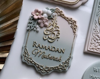 May include: A decorative plaque with a light blue and pink ombre background. The plaque has a gold ornate border and features the text "Ramadan Mubarak" in gold lettering. The plaque is decorated with a cluster of pink and white flowers and green leaves.