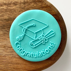 May include: A turquoise blue silicone stamp with a graduation cap and diploma design. The text "Congratulations!" is printed around the edge of the stamp.