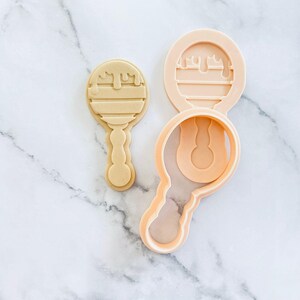 May include: A light beige cookie cutter and a peach-coloured cookie cutter set. The cookie cutter set is in the shape of a baby rattle with a dripping design on the top. The cookie cutter is on a white marble surface.