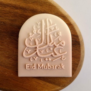 May include: A white and pink arch-shaped ceramic tile with Arabic calligraphy and the text "Eid Mubarak".