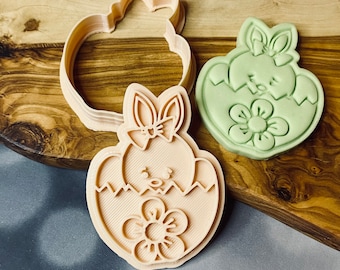 Op de afbeelding: Een plastic koekjesvormpje in de vorm van een kuiken dat uit een ei komt met een strik op zijn kop en een bloem in het ei. De vormpje is lichtbruin en heeft een gedetailleerd ontwerp.