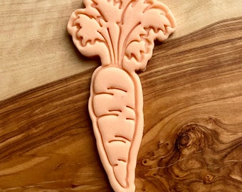 May include: A pink cookie cutter shaped like a carrot with green leafy tops. The cookie cutter is on a wooden surface.