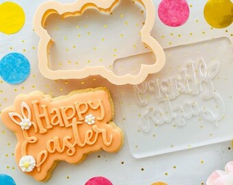 May include: A pink cookie with the words "Happy Easter" decorated with white icing flowers and bunny ears. The cookie is next to a clear plastic cookie cutter with the same words and a bunny ear design.
