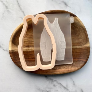 May include: A light pink plastic cookie cutter shaped like a pair of overalls. The cookie cutter is on a wooden surface next to a clear silicone mold of the same design.