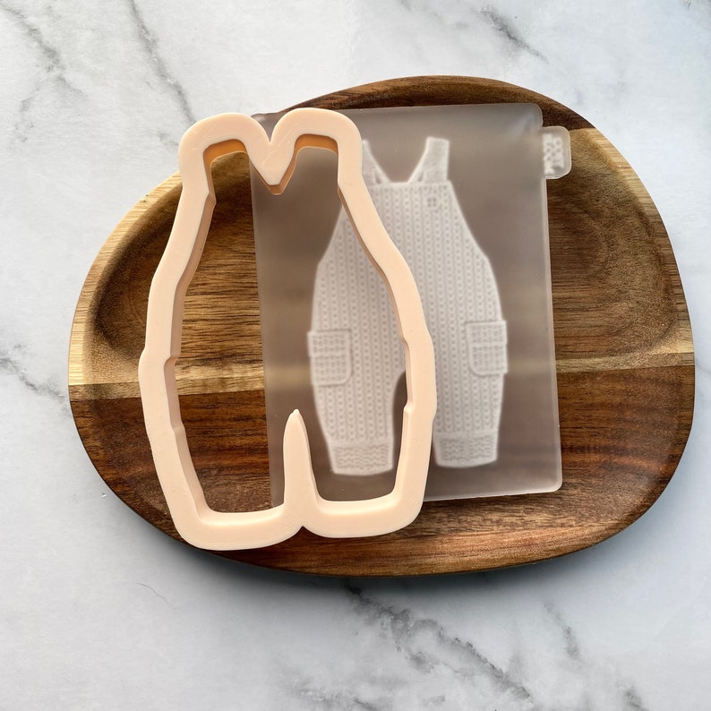May include: A light pink plastic cookie cutter shaped like a pair of overalls. The cookie cutter is on a wooden surface next to a clear silicone mold of the same design.