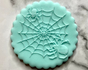 May include: A light blue cookie with a white spider web design and two spiders. The cookie is decorated with a scalloped edge.