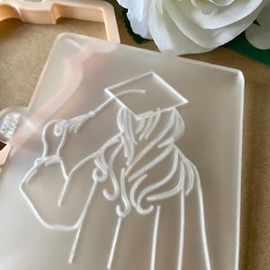 May include: A clear plastic cookie cutter with a raised design of a person wearing a graduation cap and gown. The person is holding the cap in their hand.