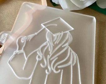 May include: A clear plastic cookie cutter with a raised design of a person wearing a graduation cap and gown. The person is holding the cap in their hand.