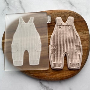 May include: A clear silicone mold with a raised design of a knitted baby overall. The mold is on a wooden surface.