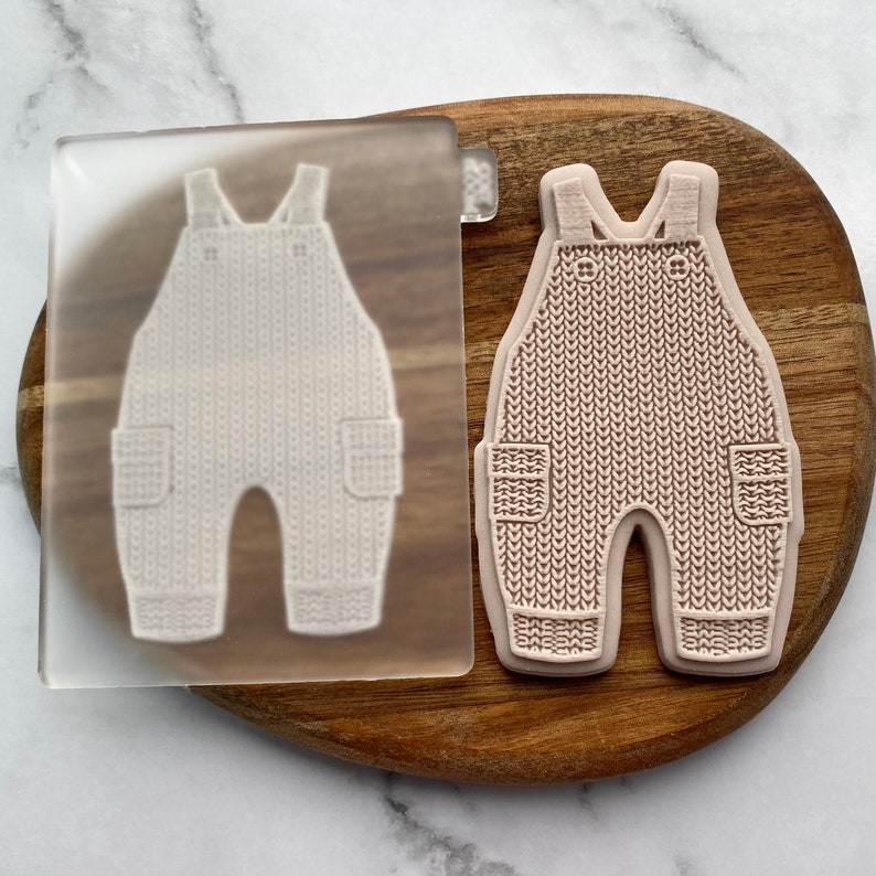 May include: A clear silicone mold with a raised design of a knitted baby overall. The mold is on a wooden surface.