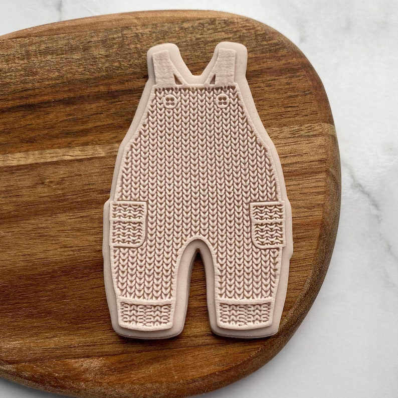 May include: A pink silicone mold shaped like a pair of knitted overalls with pockets. The mold is designed for baking and decorating.