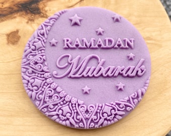 May include: A round, lavender-colored Ramadan Mubarak stamp. The design features a crescent moon with intricate patterns, stars, and the words "RAMADAN Mubarak" in elegant script. The stamp is set on a light wooden surface.