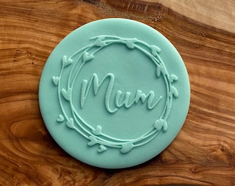 May include: A light green round cookie cutter with a wreath of leaves and hearts around the word "Mum" in the center.