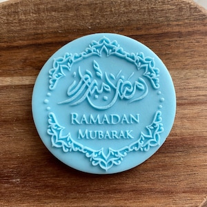 Puede incluir: Un sello de silicona azul claro con un borde de diseño floral intrincado. El sello presenta la caligrafía árabe para "Ramadan Mubarak" en el centro.