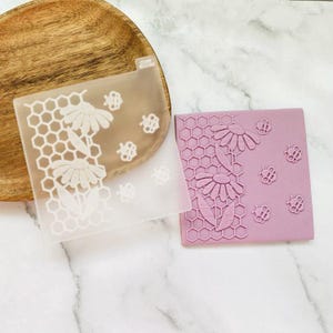 May include: Two clear silicone stamps with a honeycomb and daisy design. The stamps are on a white marble surface. The stamp on the left is clear and the stamp on the right is a light purple colour.