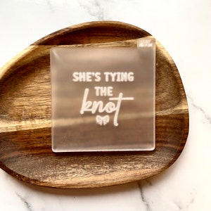 May include: A clear acrylic sign with the text "She's tying the knot" in white lettering. A small bow is below the text.
