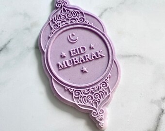May include: Purple plastic stamp with an ornate design and the text "Eid Mubarak" inside a circle. A crescent moon and two stars are also included in the design.