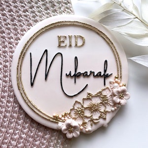 May include: A pink round cake decorated with gold glitter, gold trim, and pink flowers. The cake says "Eid Mubarak" in black script.