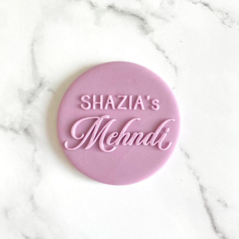 The image shows a personalized fondant embosser or stamp, for creating custom decorations on baked goods for a Mehndi ceremony. It features the custom text above the word "Mehndi" in a script font. These embossers are used to imprint designs and text onto fondant, icing, or dough, adding a personalized touch to cakes, cupcakes, and cookies for special occasions like weddings and Mehndi celebrations.