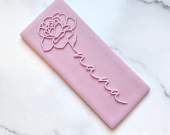 Puede incluir: Una galleta rectangular, de color lila claro, con un diseño floral y la palabra "nana" en cursiva. La flor está en la parte superior, con el tallo que conduce a la palabra. La galleta es de un color pastel suave.