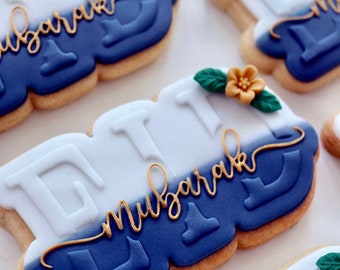 May include: Decorated sugar cookies with blue and white icing. The cookies are shaped like the word "Eid" and have the words "Eid Mubarak" written in gold icing. The cookies are decorated with small green leaves and gold flowers.