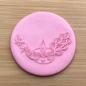 Puede incluir: Un sello de silicona rosa con un diseño blanco de una diya, una lámpara de aceite tradicional india, con un diseño de pavo real a cada lado.
