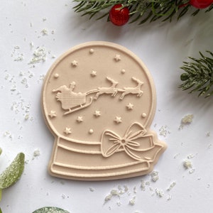 Op de afbeelding: Beige koekjesvorm in de vorm van een sneeuwbol met een kerstthema. Het ontwerp toont de kerstman in een slee getrokken door rendieren, sterren en een decoratieve strik. Feestelijk groen en decoratieve accenten omringen de vorm.