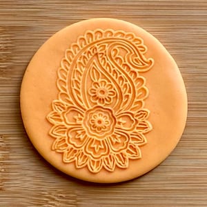 Peut inclure: Un tampon en silicone rond de couleur orange avec un motif paisley complexe. Le motif présente une fleur centrale avec un motif détaillé.